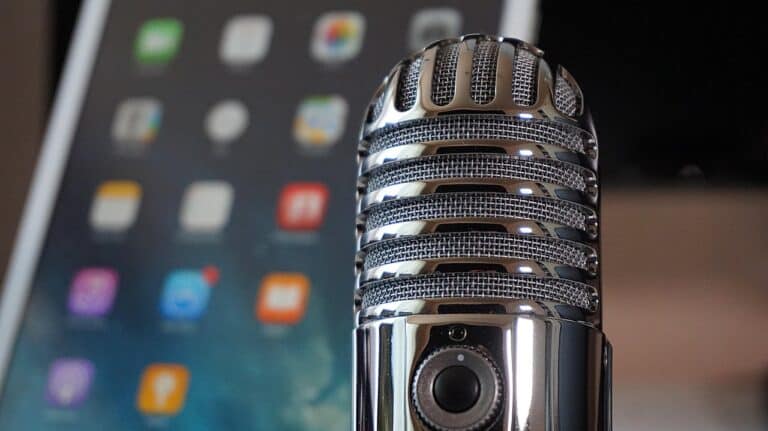 An extreme close-up of a silver, retro-style condenser microphone with a mesh grille. The background is blurred, showing a tablet screen displaying colorful app icons.